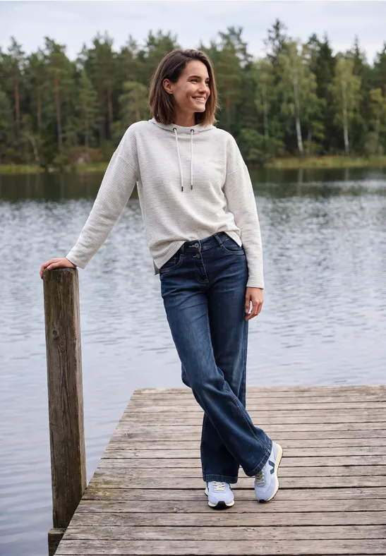 Frau in weißem Hoodie und Jeans lehnt entspannt auf einem Holzsteg am ruhigen Seeufer, umgeben von Wald.