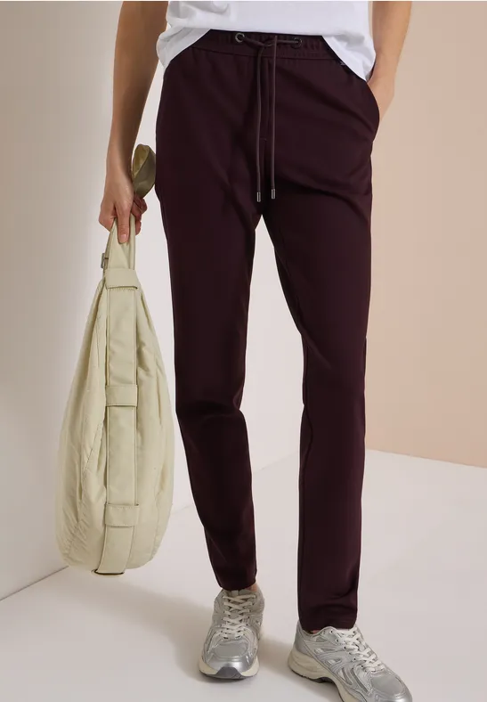 Person trägt eine weiße Bluse, bordeauxrote Hose, graue Sneaker und hält eine hellbeige Umhängetasche.