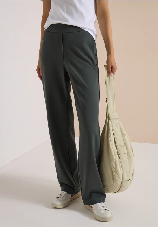 Femme en baskets blanches, pantalon noir ample et sac beige sur fond neutre