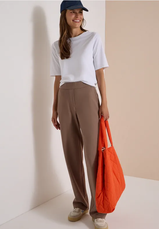 Eine junge Frau in weißem T-Shirt, brauner Hose, blauer Kappe hält eine orange Tasche.