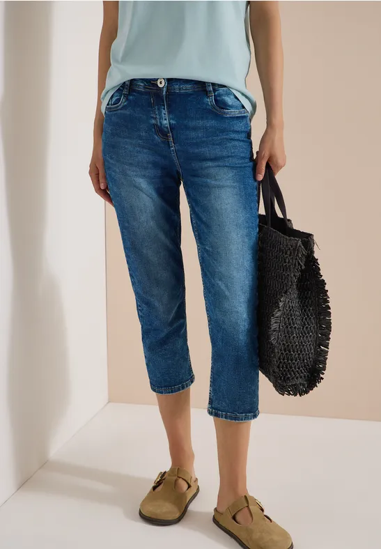 Eine Person trägt blaue Capri-Jeans, khakigrüne Sandalen und hält eine schwarze geflochtene Tasche.