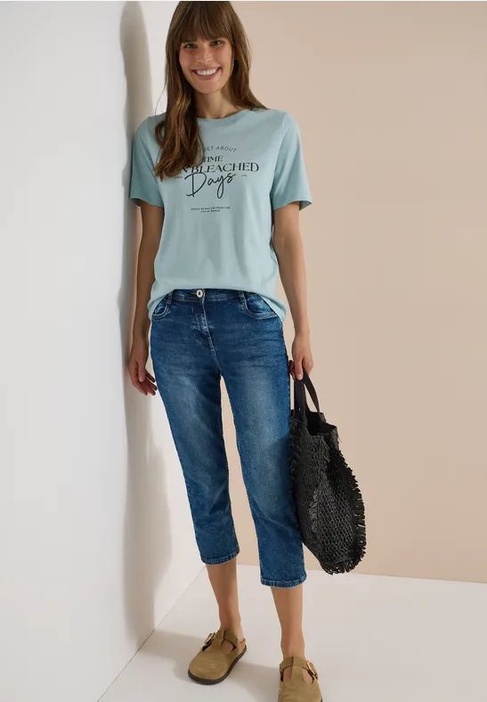 Frau in hellblauem T-Shirt, lässigen Jeans, grünen Slippern und schwarzer Korbtasche vor zweifarbigem Hintergrund.