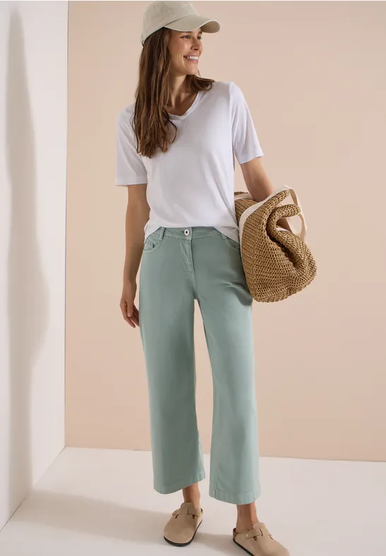 Eine Frau in weißem T-Shirt, mintgrünen Culottes, beiger Cap und Strohtasche, die seitlich lächelnd posiert.
