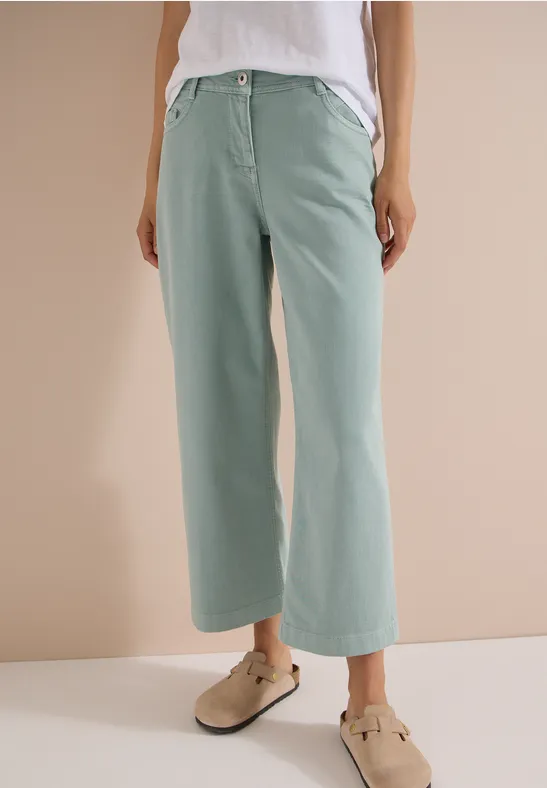 Frau trägt weite, pastellgrüne Hosen und beige Clogs vor neutralem Hintergrund.