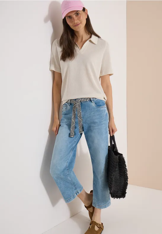 Frau lehnt an Wand, trägt weite Jeans, helles Polo, Sandalen und schwarze Häkel-Tasche.