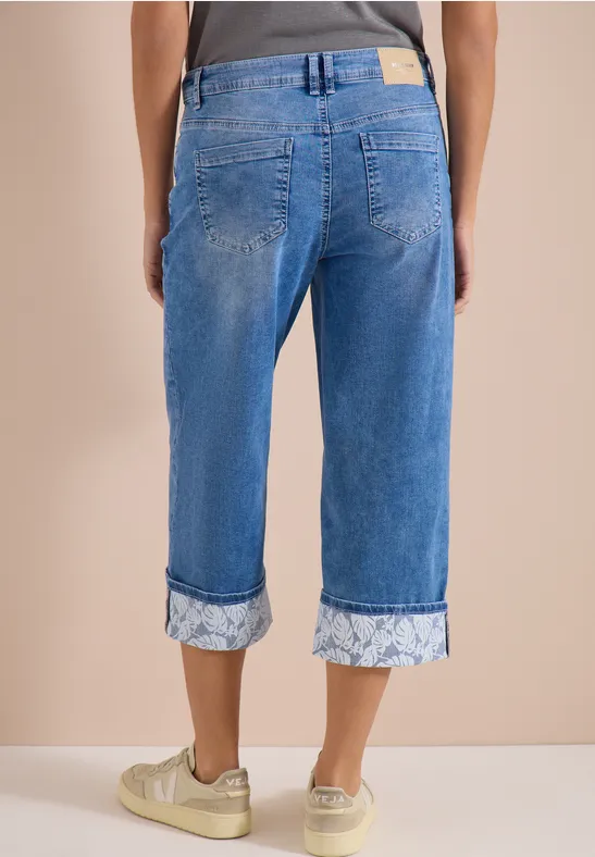 Rückansicht: Person trägt hellblaue, umgeschlagene Capri-Jeans mit floralem Muster und weiße Sneaker mit Goldakzenten.