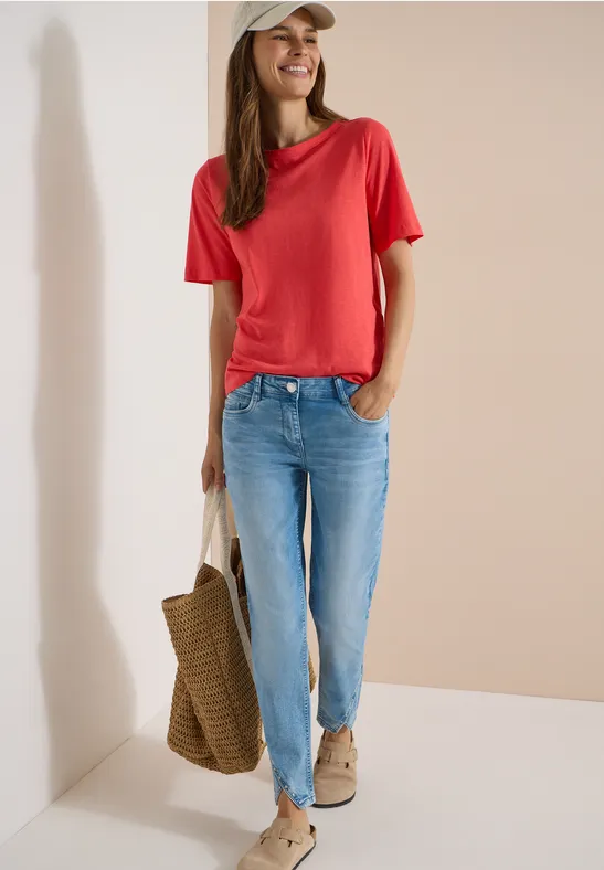Frau trägt rotes T-Shirt, blaue Jeans, bequeme Schuhe und hält eine Strohtasche vor hellem Hintergrund.