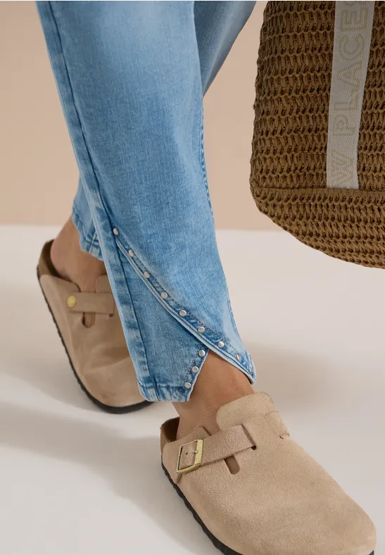 Beige Wildleder-Clogs mit goldener Schnalle, kombiniert mit hellblauen Jeans mit Knopfschlitz und geflochtener Tasche.