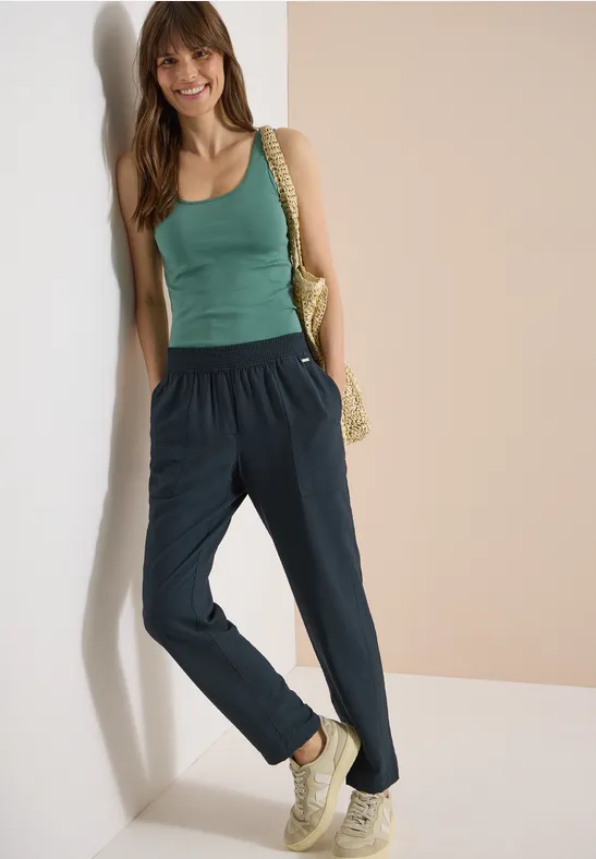 Frau lehnt an Wand, trägt grünes Top, blaue Stoffhose, Sneakers und Korbtasche.