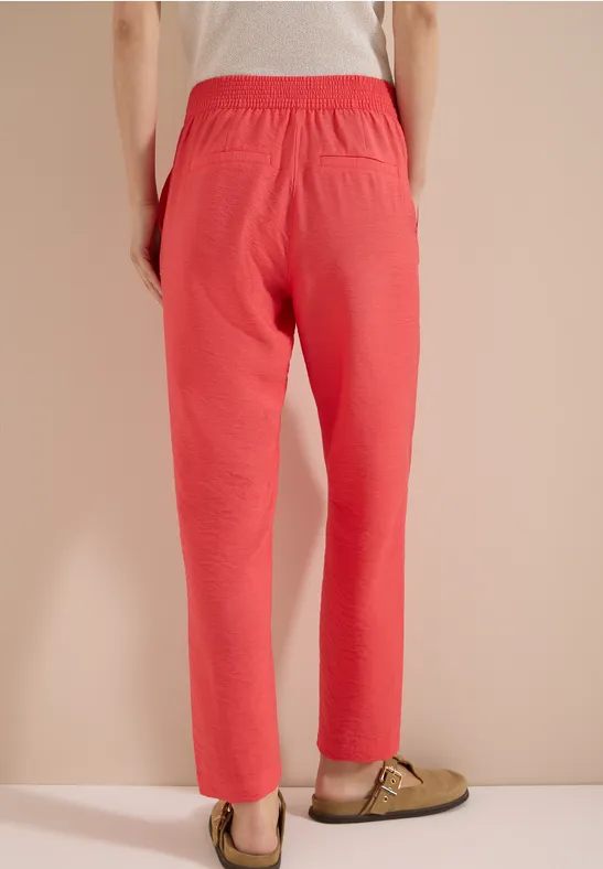 Person steht vor beiger Wand und trägt eine korallrote Hose mit grünen Pantoffeln.