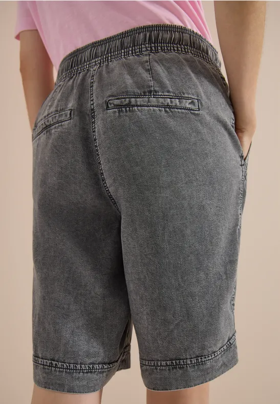 Une personne porte un bermuda en denim gris avec taille élastiquée, poches arrière et latérales, mains dans les poches.