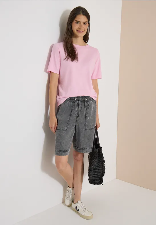 Une femme porte un t-shirt rose, un short bermuda gris, des baskets blanches et un sac noir.