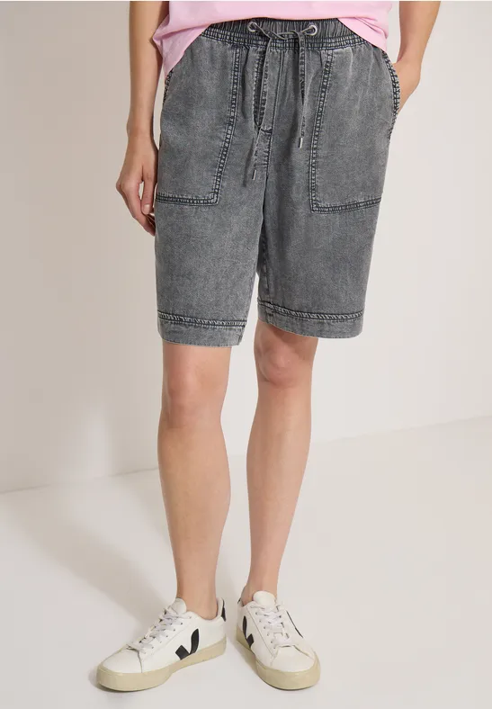 Personne portant un short en jean gris avec poches et baskets blanches sur fond neutre.