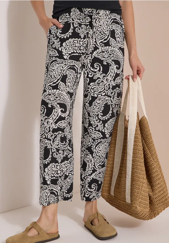 Personne porte un pantalon de détente noir et blanc à motif cachemire, des sandales et tient un grand sac en paille.