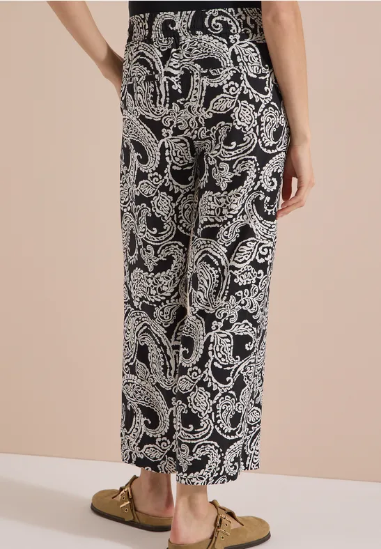 Pantalon large noir 7/8 à motif paisley blanc, porté avec des mocassins dorés sur fond beige.