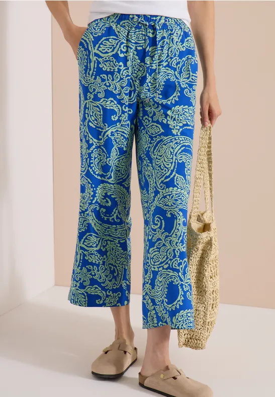 Blaue Hose mit weitem Bein und grünen Paisley-Muster, kombiniert mit Basttasche und goldenen Schuhen.