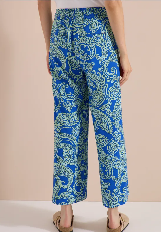 Rückansicht einer Person in weiten Hosen mit blau-grünem Paisley-Muster und flachen Sandalen.