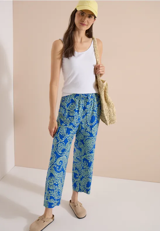 Frau in weißem Top und blauen, gemusterten Culotte-Hosen mit Strohtasche vor hellem Hintergrund.