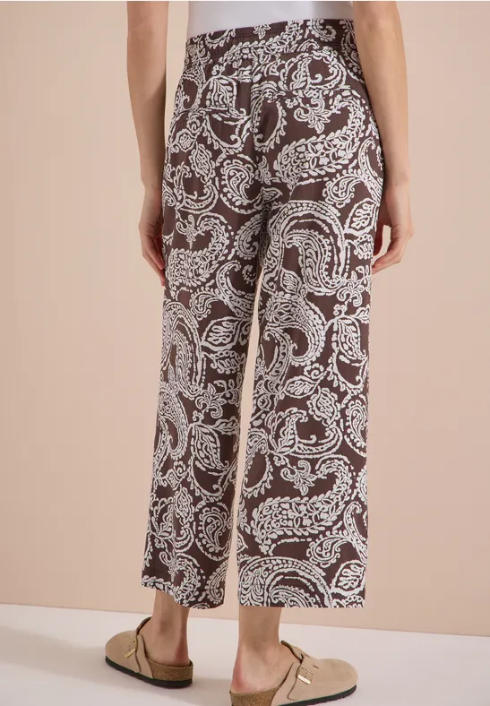 Rückenansicht einer Person in braunen, knöchellangen Culottes mit weißem Paisley-Muster und beige Loafers.
