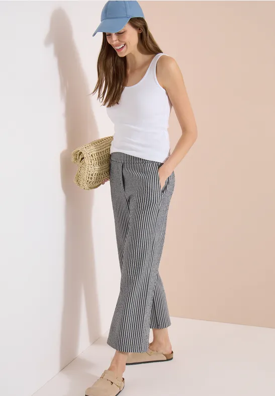 Frau in weißem Top, grauen, karrierten Hosen, blauem Cap und Strohtasche lehnt lässig an der Wand.