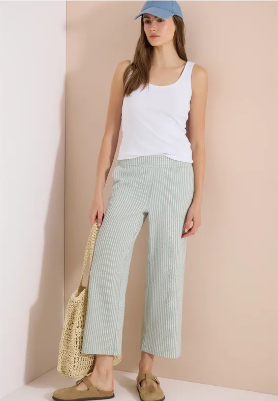 Vrouw in witte tanktop en mintgroene wijde cropped broek, met strooien tas voor een neutrale achtergrond.