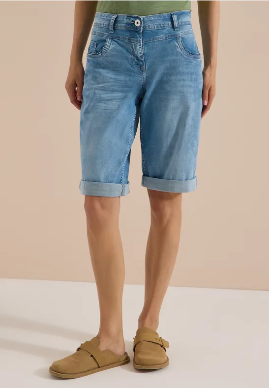 Person trägt hellblaue Jeans-Bermudashorts und beigefarbene Sandalen vor neutralem Hintergrund.