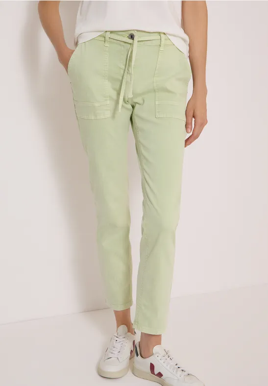 Frau trägt helle grüne Chinohose, weißes Shirt und weiße Sneaker, frontal stehend, Hände in Taschen.