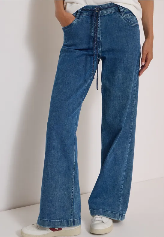 Personne portant un jean bleu taille haute, coupe évasée et baskets blanches.