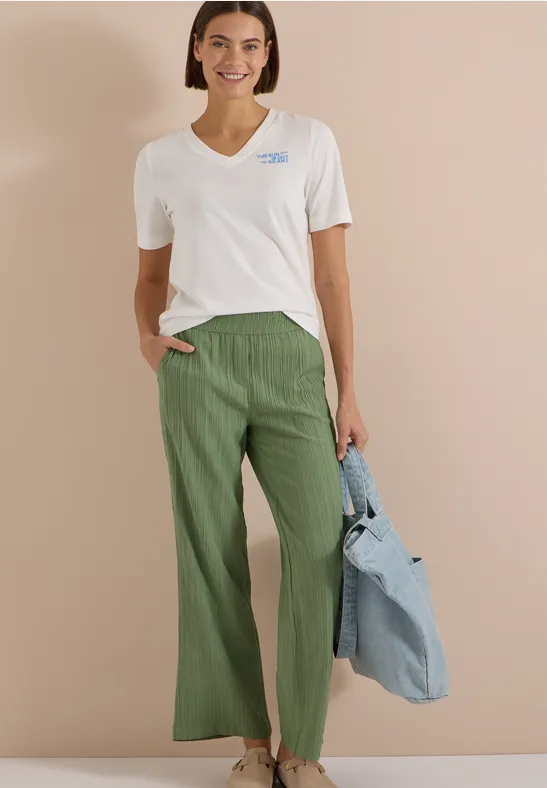 Femme en t-shirt blanc, pantalon vert ample et sac rayé devant un fond neutre.