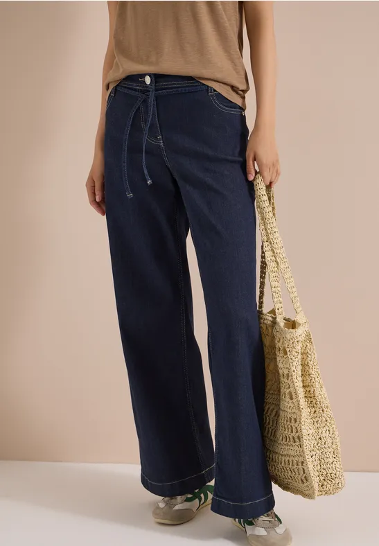 Frau trägt weite, dunkle Jeans, beige Top, Sneakers und eine gehäkelte Tasche vor neutralem Hintergrund