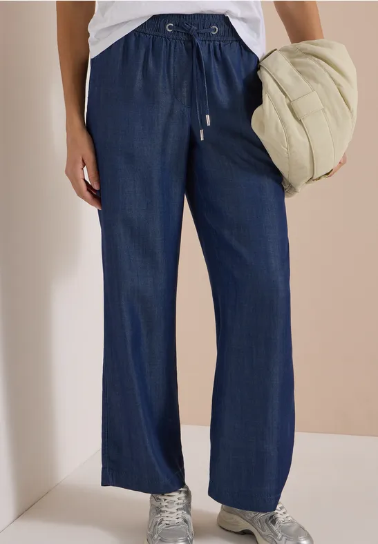 Pantalon jean bleu foncé, taille coulissée et coupe droite, porté avec des baskets et une doudoune claire.