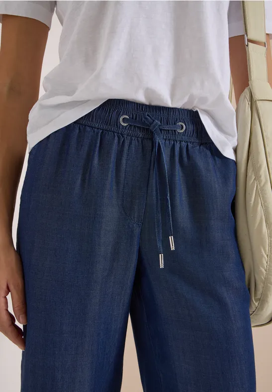 Personne en t-shirt blanc, pantalon bleu foncé à cordon de serrage et sac à épaule clair.