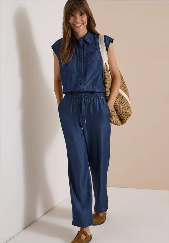 Frau in ärmellosem Jeans-Jumpsuit mit Strohtasche und Sandalen, die lächelnd posiert.