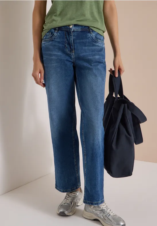 Frau in blauen Jeans mit weitem Bein, grünes Top, graue Sneaker, hält eine dunkelblaue Tasche.