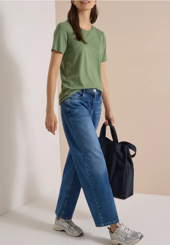 Eine Person trägt grünes T-Shirt, weite blaue Jeans, weiße Sneaker und hält eine dunkelblaue Tasche.