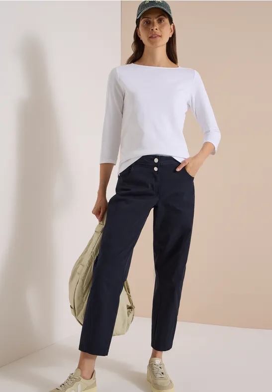 Frau trägt weißes Shirt, dunkelblaue Hose, Sneaker und Cap, hält Jacke lässig über die Schulter.