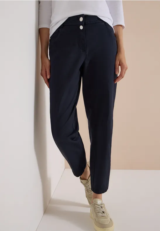 Person lehnt an Wand, trägt dunkelblaue, hochgeschnittene Hose mit Knopfleiste und beige Sneaker.