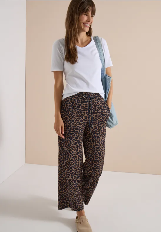 Femme souriante en t-shirt blanc, pantalon large léopard, baskets et sac d’épaule.