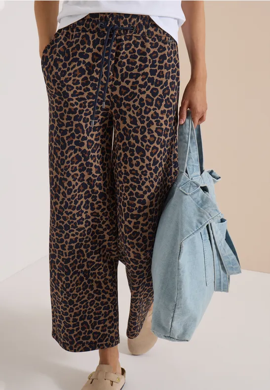 Une personne porte un pantalon léopard ample, des baskets claires et tient une veste en jean devant un fond neutre.