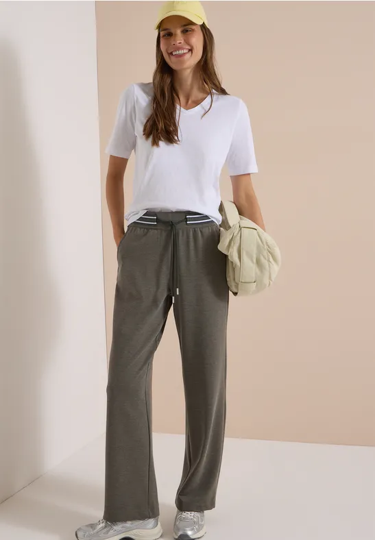 Frau trägt weißes T-Shirt, weite olivgrüne Hose, gelbe Kappe, hält Tasche und lächelt.