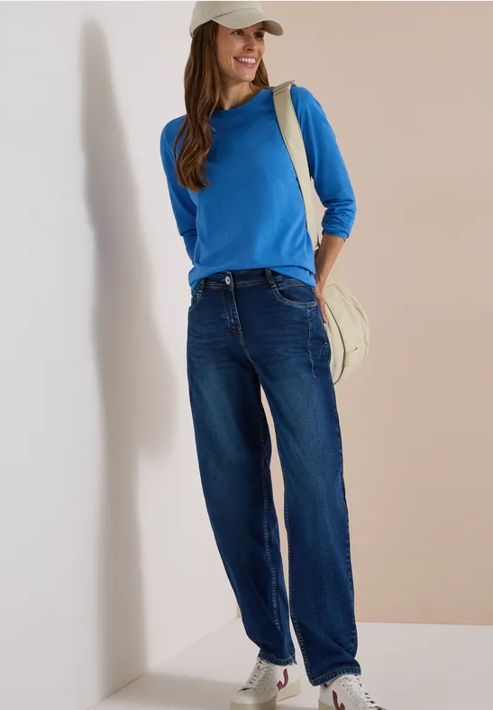 Eine junge Frau in blauem Pullover und Jeans, mit Cap und Rucksack vor weißer Wand.