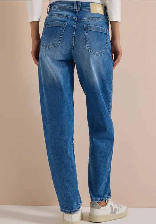 Rückansicht einer Person in locker sitzender blauer Jeans und weißen Sneakers vor beigefarbenem Hintergrund.