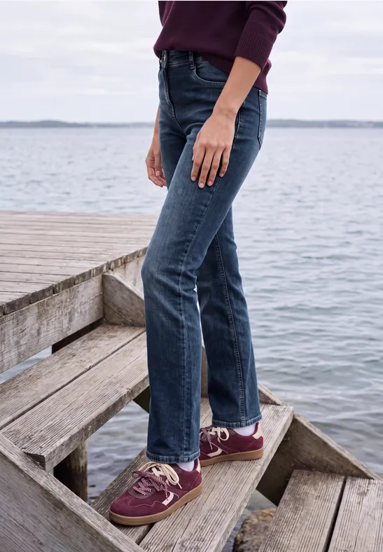 Personne en jean et baskets bordeaux debout sur des marches en bois au bord de l'eau.