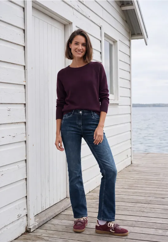 Femme en pull violet et jean, souriante sur une jetée devant une maisonnette blanche au bord de l'eau.