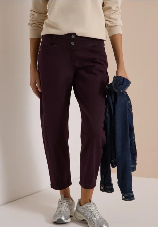 Personne porte un haut beige, un pantalon bordeaux, des baskets argentées et tient une veste en jean.