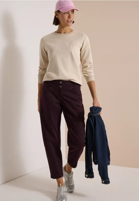Femme en sweat clair, pantalon bordeaux, casquette rose, baskets argentées, tenant une veste bleue.