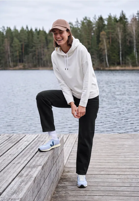 Femme souriante en sweat blanc et pantalon noir sur un ponton au bord d'un lac, forêt en arrière-plan.