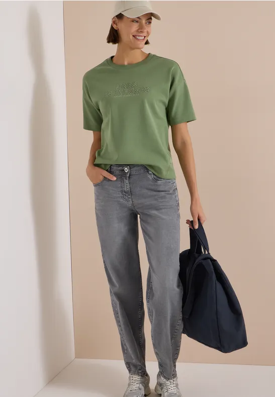 Frau in grünem T-Shirt, grauer Jeans und Cap, mit großer dunkelblauer Tasche vor beigem Hintergrund.