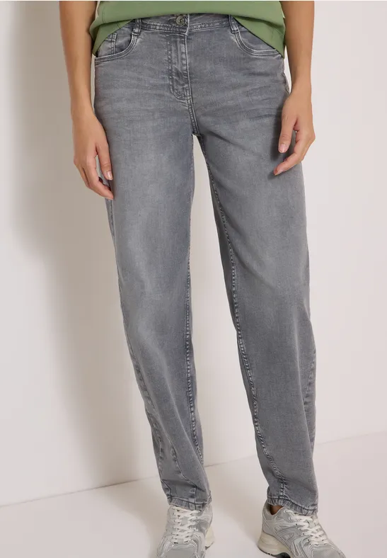 Person trägt gerade geschnittene graue Jeans und Sneaker, vor hellem Hintergrund stehend.