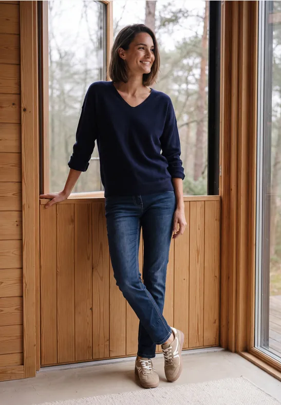 Une femme s'appuie sur un cadre de fenêtre en bois, porte un jean et un pull marine et sourit.